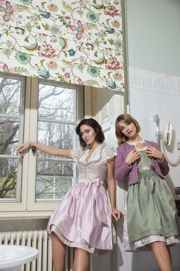 Women wearing Kruger Mini Dirndl dresses, one in pink with ornamental bodice and skirt, styled in a vintage setting.